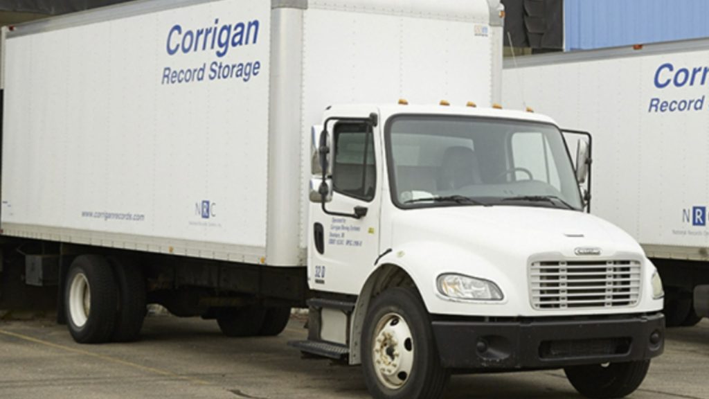 White delivery truck with "Corrigan Record Storage" logo is parked beside similar trucks. Overcast lighting suggests a business setting.