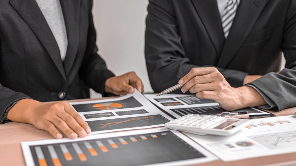 Two people in business attire review financial charts and graphs on paper, with a calculator placed on the table.